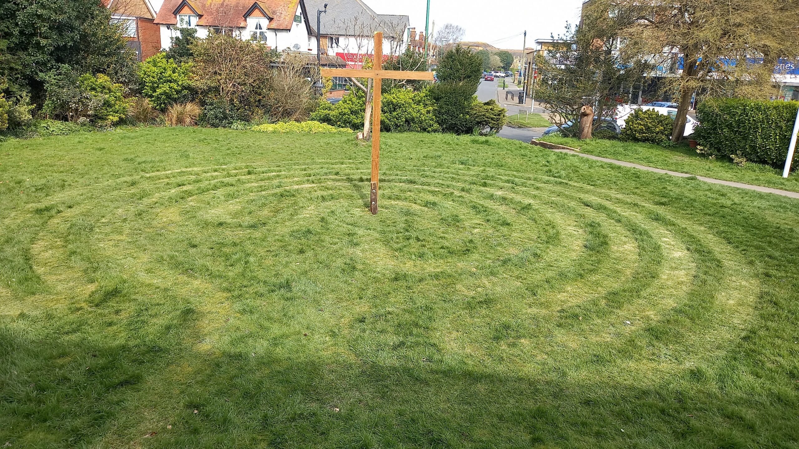 A grassy area featuring a wooden cross at centre surrounded by concentric circular patterns mowed into the grass. Shops, houses and trees are visible in the background, indicating a community setting.